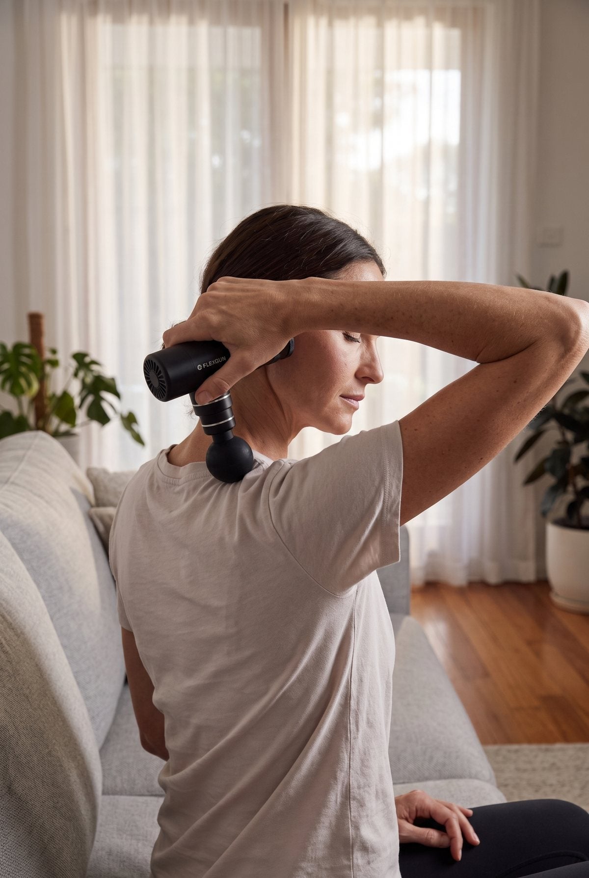 Woman on a couch reaching over her shoulder with the Flexgun mini onto the top of her upper back